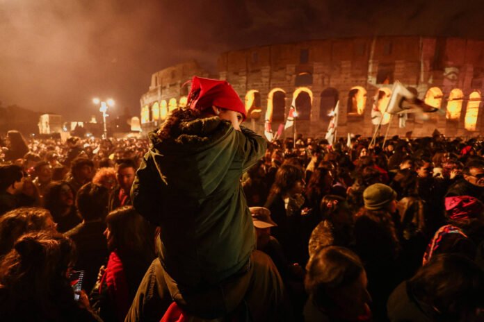 Grande grupo de pessoas aglomeradas à noite em frente ao Coliseu iluminado em Roma. Uma criança com gorro vermelho está no ombro de um adulto, e várias bandeiras são agitadas na multidão.