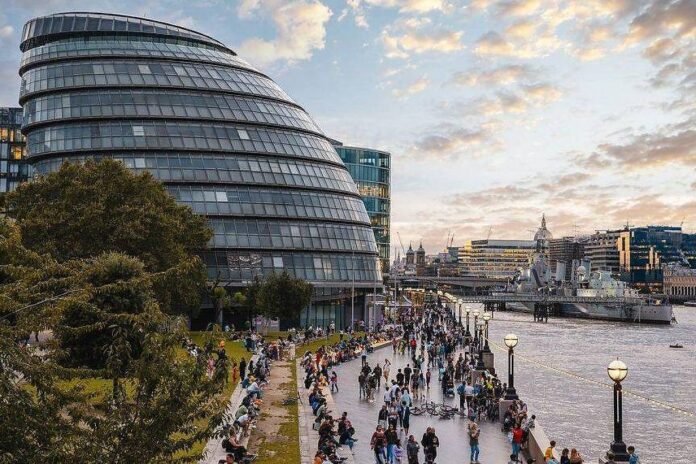 Multidão caminha e senta-se ao longo da margem do rio Tâmisa em Londres, com o edifício curvo e moderno conhecido como City Hall à esquerda. Ao fundo, navio de guerra está ancorado no rio, e o céu apresenta nuvens ao entardecer.