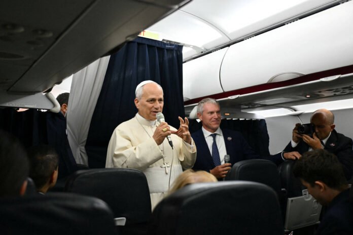 Papa Francisco fala ao microfone dentro de avião, acompanhado por homem de terno segurando outro microfone. Pessoas sentadas observam, e fotógrafo registra cena ao fundo.