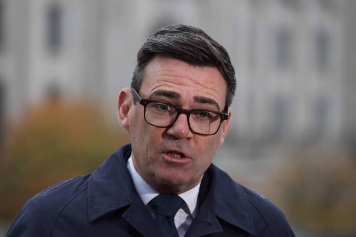 Homem de meia-idade com cabelo curto e escuro, usando óculos de armação preta, terno azul escuro, camisa branca e gravata azul, fala em ambiente externo com fundo desfocado de edifício e vegetação.