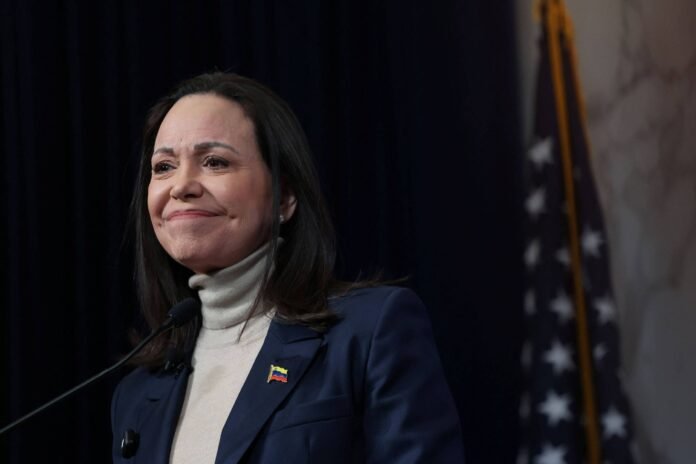 Mulher de cabelos escuros usa paletó azul e blusa de gola alta branca, com broche da bandeira da Venezuela, fala em púlpito. Ao fundo, bandeira dos Estados Unidos desfocada.