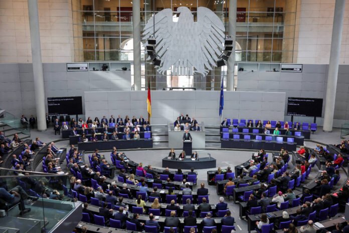 Plenário do Bundestag em Berlim com dezenas de parlamentares sentados em cadeiras azuis dispostas em semicírculo. Orador fala em púlpito central sob grande águia prateada na parede. Bandeiras da Alemanha e da União Europeia estão ao fundo.