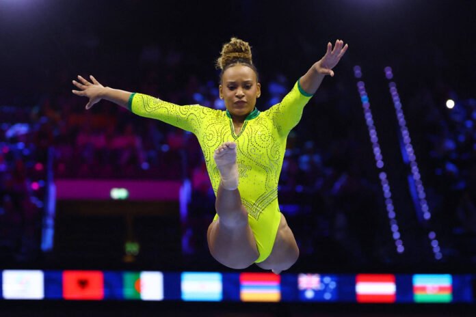 Ginasta feminina em collant amarelo vibrante executa salto com pernas estendidas e braços abertos em arena escura com público ao fundo.