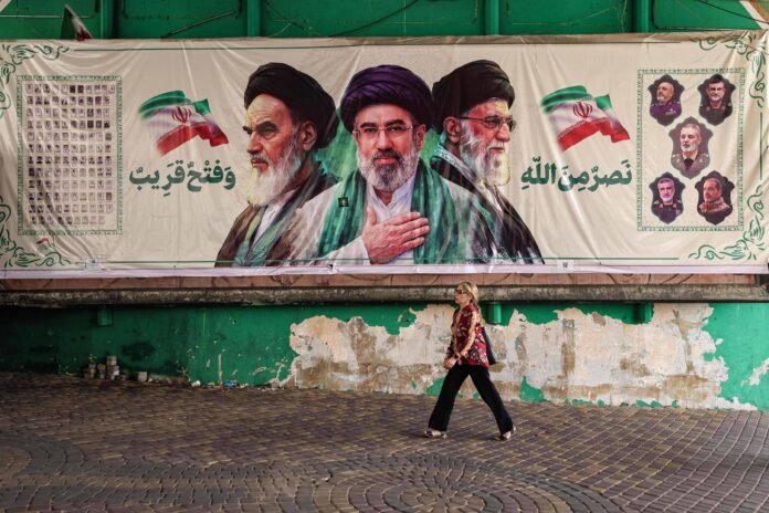 Mural grande exibe três retratos de líderes iranianos com turbantes pretos, entre duas bandeiras do Irã e textos em persa. Uma mulher caminha em primeiro plano sobre calçada de pedras, com parede verde e descascada ao fundo.