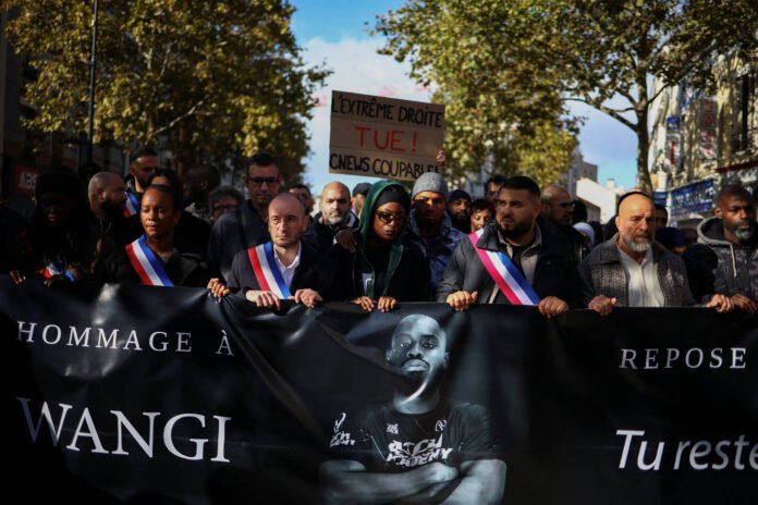 Grupo de manifestantes caminha em rua arborizada segurando faixa preta com foto de Wangi e texto em francês homenageando-o. Alguns usam faixas tricolores. Ao fundo, cartaz denuncia extrema direita.